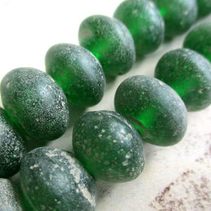 May include: Close-up of a strand of oval-shaped, translucent green glass beads. The beads have a textured, weathered appearance with white speckling, suggesting an aged or vintage aesthetic. The beads are arranged in a diagonal row.