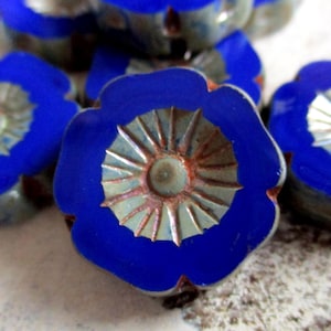 Op de afbeelding: Een verzameling blauwe en zilveren glazen knopen in de vorm van een bloem met een verhoogd centraal ontwerp. De knopen hebben een metallic glans en een blauwe rand.
