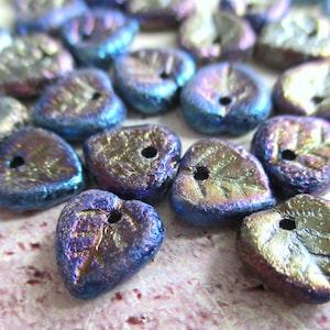 Può includere: Una collezione di perline di vetro a forma di cuore di colore blu iridescente e oro con un design a foglia. Le perline hanno una superficie testurizzata e un foro al centro per l'infilaggio.