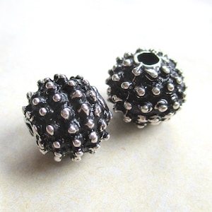 Peut inclure: Deux perles sphériques avec une finition sombre et oxydée et une surface texturée comportant de nombreuses bosses argentées polies en relief. Chaque perle possède un trou central, créant un contraste marqué entre les zones sombres en creux et les saillies métalliques brillantes sur toute la surface extérieure.