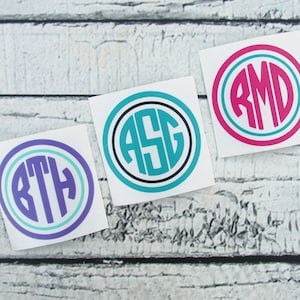 May include: Three circular monogram stickers with white backgrounds. Each sticker features a different three-letter monogram in a bold, sans-serif font. The monograms are in teal, purple, and pink, with coordinating accent colors and black outlines. The stickers are on a white-washed wooden surface.