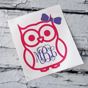 May include: A pink owl decal with a purple bow and a monogram in the center. The owl has large eyes and a cute design. The monogram features the letters MBL in a stylish font. The decal is on a white background.