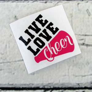 May include: A white square sticker with the words "LIVE LOVE" in bold black letters above a pink megaphone shape with the word "cheer" in white script. The sticker is on a white brick background.