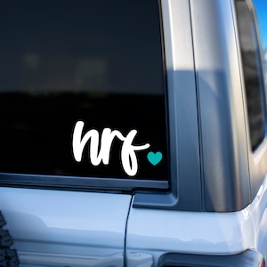 May include: White vinyl decal with the text "hrf" and a small teal heart on a black background. The decal is applied to the back window of a silver car.