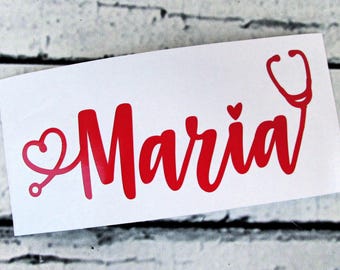 May include: A red vinyl decal featuring the name Maria in a cursive script font. The letter M is integrated with a stethoscope design that forms a heart shape on the left side, and a small red heart is positioned above the letter i.