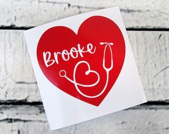 May include: A square white decal featuring a large red heart. Inside the heart, the name Brooke is written in white script lettering, accompanied by a white line drawing of a stethoscope that forms a smaller heart shape at the bottom.
