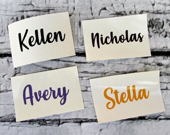 May include: Four rectangular adhesive vinyl decals featuring custom names in a cursive script font. The top left decal displays the name Kellen in black, top right shows Nicholas in black, bottom left shows Avery in purple, and bottom right shows Stella in orange.