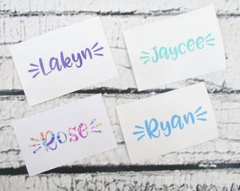 May include: Four rectangular white stickers featuring custom names in various colors and fonts. The names Lakyn in purple, Jaycee in teal, Rose in a multi-colored floral pattern, and Ryan in blue are each flanked by small decorative lines.