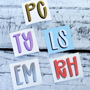May include: Five square stickers with different letter combinations: "PC" in gold and black, "TY" in purple, "LS" in white on a blue background, "EM" in gray, and "RH" in red. The stickers are arranged on a white and gray wooden surface.
