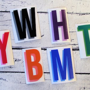 May include: A collection of colorful letter stickers. The letters are in various colors, including red, orange, blue, purple, green, and black, each with a shadow effect. The stickers are on a white background.