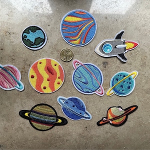 Op de afbeelding: Een set van tien geborduurde strijkpatches met planeten en een raket. De patches zijn kleurrijk en hebben verschillende ontwerpen, waaronder een blauwe en groene planeet met ringen, een rode en gele planeet met ringen, een blauwe en witte planeet met ringen, een gele en oranje planeet met ringen, een blauwe en witte planeet met ringen, een blauwe en witte planeet met ringen, een blauwe en witte planeet met ringen, een blauwe en witte planeet met ringen, een blauwe en witte planeet met ringen en een grijze raket met een blauw raam en rode vlammen.