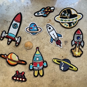Könnte beinhalten: Eine Sammlung von zehn gestickten Bügeletiketten mit Weltraummotiven. Die Patches zeigen Raketen, Planeten und Sterne. Die Patches sind in verschiedenen Farben und Größen erhältlich. Die Patches sind perfekt, um Kleidung, Taschen oder anderen Gegenständen einen Hauch von Verspieltheit zu verleihen.