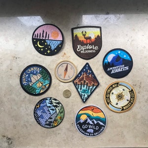 May include: Nine embroidered iron-on patches with various nature and adventure themes. The patches feature a deer, a moon, a compass, a campfire, a mountain, a tree, and text that reads "Explore Wilderness", "Adventure Awaits", "Happy Camper", "Not All Those Who Wander Are Lost", and "Go Wild Adventure Is Calling".
