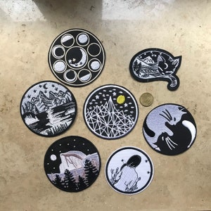 Op de afbeelding: Een verzameling geborduurde patches met hemelse en natuurlijke thema's. Ontwerpen omvatten maanfasen, berglandschappen, een katten silhouet met een berglandschap en een yin-yang katten ontwerp. De patches zijn zwart en wit met gele accenten.