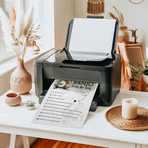 K&ouml;nnte beinhalten: Ein schwarzer Drucker druckt ein Dokument mit dem Titel "Banjo Facts for Kids". Das Dokument enth&auml;lt eine Banjo-Illustration und Text. Der Drucker steht auf einem wei&szlig;en Tisch, im Hintergrund eine Vase mit Pampasgras und andere dekorative Gegenst&auml;nde.