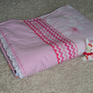 Puede incluir: Una colcha de retazos rosa y blanca con un ribete de encaje blanco. La colcha está doblada por la mitad y un pequeño animal de peluche está encima.