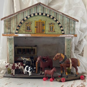 Könnte beinhalten: Ein Vintage-Spielzeugstall aus Holz mit grün-braun lackierter Außenseite. Im Inneren befinden sich mehrere Holztiere, darunter Kühe und Pferde. Die Pferde sind auf Rädern. Die Scheune hat eine bemalte Tür und Fenster.