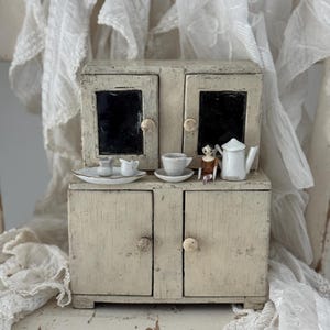 Puede incluir: Una alacena de madera en miniatura de color blanco roto con cuatro puertas y dos pequeños pomos en cada nivel. La alacena muestra un pequeño juego de té de cerámica blanca que incluye una tetera, una taza con plato, una lechera y una pequeña figura.