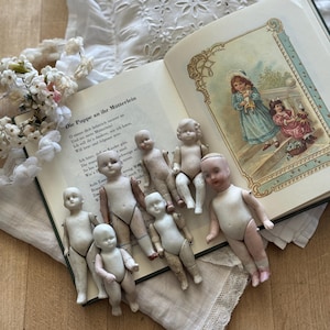 Pode incluir: Uma coleção de bonecas antigas de porcelana de vários tamanhos, dispostas em um livro vintage com uma ilustração. As bonecas são em sua maioria brancas com traços pintados, algumas mostrando sinais de idade. A cena é montada em uma superfície de madeira com um pano com borda de renda.