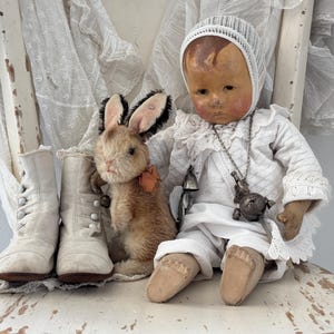 Könnte beinhalten: Eine Porzellanpuppe in einem weißen Kleid und einer Haube sitzt auf einem weißen Stuhl mit einem Plüsch-Hasen aus braunem Stoff. Die Puppe hat einen weißen Spitzenbesatz an den Ärmeln des Kleides und eine silberne Halskette mit einem Anhänger.