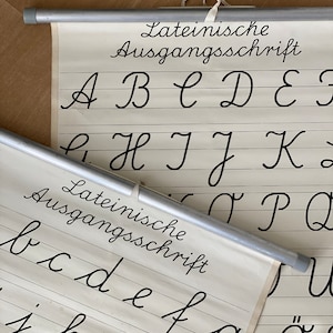 May include: Two vintage cursive handwriting charts with metal rods. The charts display the alphabet in uppercase and lowercase, with the words "Lateinische Ausgangsschrift" at the top. The letters are in black ink on a cream-coloured background.
