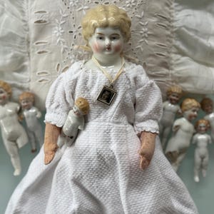 Op de afbeelding: Een vintage porseleinen pop met blond haar en een witte jurk, die een kleinere pop vasthoudt. Een hanger met een foto hangt om haar nek. Verschillende kleine porseleinen poppen omringen de hoofdpop, met een witte kanten achtergrond.