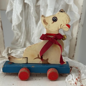 Può includere: Un giocattolo vintage in legno a forma di cane. Il cane ha un corpo color crema, un fiocco rosso e una piccola campana dorata. È montato su una base blu con ruote rosse. Il giocattolo ha un'estetica classica e antica.