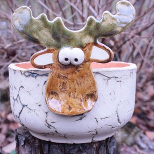Könnte beinhalten: Ein skurriler Keramik-Pflanztopf mit einem Elch-Design. Der Elch hat große, ausdrucksstarke Augen und ein Geweih. Der Topf ist weiß mit einer rissigen Textur und einem pfirsichfarbenen Innenraum. Der Elch ist braun und grün.