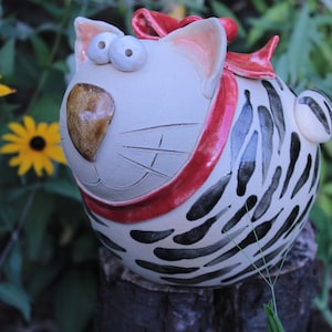 Könnte beinhalten: Eine runde Keramik-Katzenfigur mit cremefarbenem Körper, schwarzen Streifen und einem roten Halsband. Die Katze hat große, weit auseinanderstehende Augen, eine braune Nase und eine rote Schleife auf dem Kopf. Sie hat einen kleinen Schwanz.