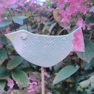 Könnte beinhalten: Ein Keramik-Vogel-Gartenstecker mit einem cremeweißen Körper, einem hellblauen geometrischen Prägemuster, einem kleinen gelben Schnabel, einem einzelnen dunklen Auge und einem rosafarbenen Schwanz, montiert auf einem dünnen, hellen Holzstab.