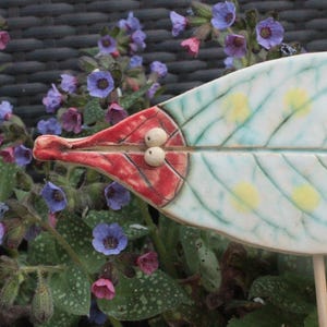 Könnte beinhalten: Ein dekorativer Gartenstecker aus Keramik in Blattform. Das Blatt ist weiß mit grünen Adern und gelben Punkten. Der Stiel ist hellbraun. Das Ende ist rot mit zwei kleinen weißen Kugeln. Der Stecker ist von lila und rosa Blüten umgeben.