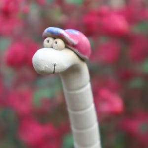 Könnte beinhalten: Eine skurrile Keramik-Wurmfigur mit weißem Körper und lächelndem Gesicht. Sie trägt eine rosa und blaue Baseballkappe. Der Hintergrund ist eine verschwommene Darstellung rosa Blüten.