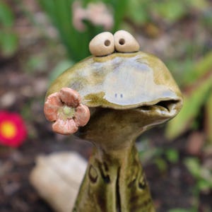 Könnte beinhalten: Keramikfroschfigur mit Blumendetail. Der Frosch ist hauptsächlich grün mit braunen Augen und einer Blume auf der Wange. Der Hintergrund ist verschwommen mit grünen und roten Blumen.