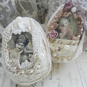 May include: Two decorative egg-shaped ornaments. One is white with a vintage portrait, lace, and pearls. The other has a similar design with a portrait, pink roses, and ribbon. The ornaments are on a vintage paper background.