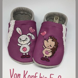 Könnte beinhalten: Ein Paar violette Lederschuhe für Babys mit gestickten Motiven eines Hasen, der Ballons hält, und eines Igels, der Ballons hält. Die Schuhe haben einen weißen Spitzenbesatz und der Text "Von Kopf bis Fuß" ist auf der Unterseite aufgedruckt.