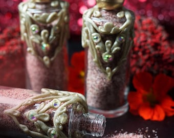 Op de afbeelding: Drie doorzichtige glazen flesjes gevuld met roze korrelig poeder. Elk flesje heeft een goudkleurige bolvormige dop en gedetailleerde gouden filigraanversieringen rond de hals, geaccentueerd met kleine, iriserende veelkleurige strass-steentjes.