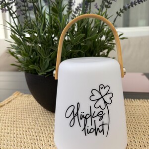 Könnte beinhalten: Eine weiße, glockenförmige, tragbare Laterne mit einem hellen Holzgriff. Auf der Vorderseite der Laterne befindet sich der schwarze Schriftzug "Glückslicht" sowie eine Grafik eines vierblättrigen Kleeblatts mit einem kleinen Herzdetail.