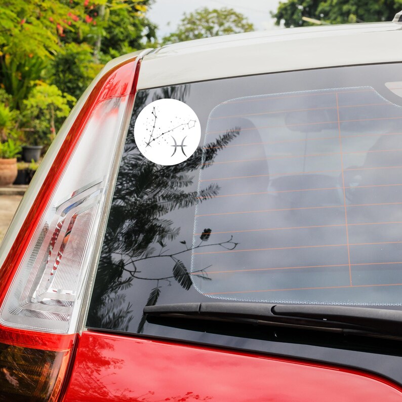 May include: A white car with a black rear window wiper. A white circle sticker with a black outline is on the rear window. The sticker features a black line drawing of the Pisces zodiac constellation with the Pisces symbol.