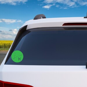 May include: A white car with a green circle sticker on the back window. The sticker features a black outline of the Pisces constellation with the Pisces symbol.