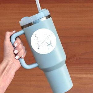 May include: A light blue travel mug with a white circle sticker featuring the Pisces zodiac constellation and symbol. The mug has a handle and a lid with a straw.