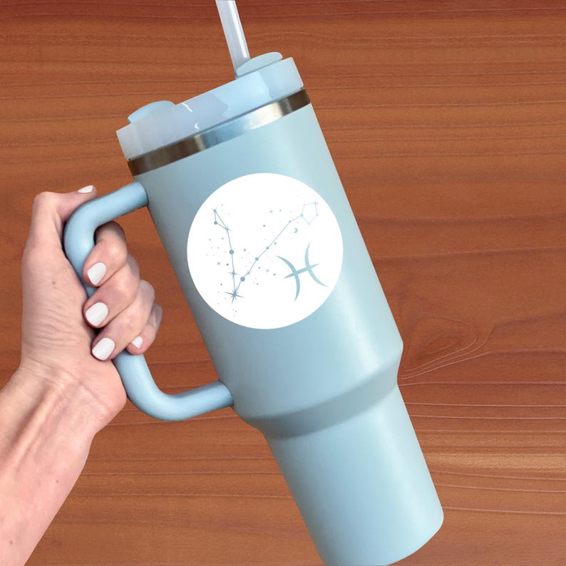 May include: A light blue travel mug with a white circle sticker featuring the Pisces zodiac constellation and symbol. The mug has a handle and a lid with a straw.