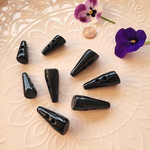 Könnte beinhalten: Eine Sammlung glänzender schwarzer Knebelknöpfe in verschiedenen Größen und Formen, die auf einem hellrosa, dekorativen Teller verstreut sind. Zwei violette Stiefmütterchen und eine Münze sind ebenfalls vorhanden und verleihen der Komposition Farbe und Detail.