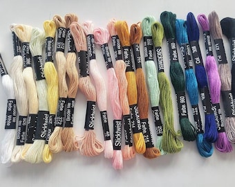 May include: A selection of embroidery threads in a range of colours, from white and yellow to pink, orange, green, blue, and purple. Each skein is labelled with a black band showing the colour number and brand.