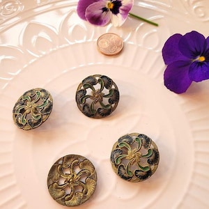 Könnte beinhalten: Vier antike Messingknöpfe mit aufwendigen floralen Designs, akzentuiert mit grüner und schwarzer Emaille. Die Knöpfe sind auf einem rosa Teller angeordnet, zusammen mit zwei violetten Stiefmütterchen und einer Münze.