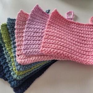 Könnte beinhalten: Ein Stapel von sieben gehäkelten Geschirrtüchern in verschiedenen Farben, darunter marineblau, türkis, grün, gelb, rosa und lila. Die Geschirrtücher sind gefaltet und übereinander gestapelt.