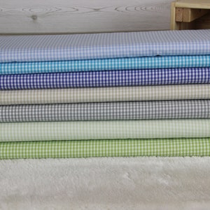 Könnte beinhalten: Ein Stapel von acht Gingham-Stoffmustern in verschiedenen Farben, darunter hellblau, türkis, lila, beige, grau und grün. Der Stoff ist ordentlich gefaltet und in einer Reihe angeordnet.