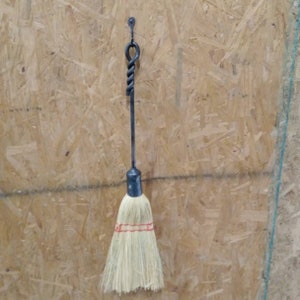 May include: A black metal fireplace tool with a twisted handle and a brush made of natural fibers. The tool is hanging on a wall.