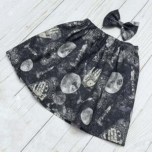 May include: A black skirt with an elastic waistband featuring a white print of ouija boards, crystal balls, candles, and celestial patterns, accompanied by a matching bow hair accessory with the same occult-inspired graphic print design.
