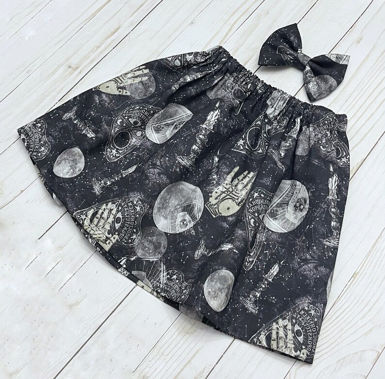 May include: A black skirt with an elastic waistband featuring a white print of ouija boards, crystal balls, candles, and celestial patterns, accompanied by a matching bow hair accessory with the same occult-inspired graphic print design.