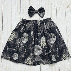 May include: A black skirt with an elastic waistband and a matching bow hair accessory. The fabric features a repeating print of white and grey occult-themed illustrations, including ouija boards, candles, moons, and hands on a dark background.